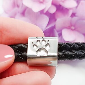 Nahaufnahme eines silbernen Anhängers mit Pfotenabdruck auf einem schwarzen geflochtenen Lederarmband, gehalten von einer Hand mit rosa Nagellack; lila Blumen im Hintergrund. Aurora Tierkrematorium