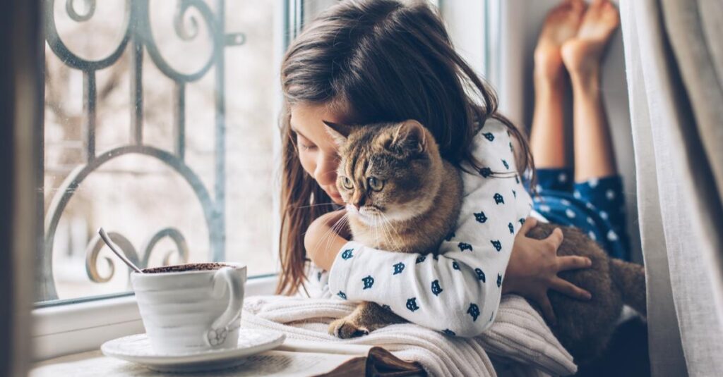 Ein junges Mädchen liegt am Fenster und umarmt eine braun getigerte Katze. Eine Tasse mit einem heißen Getränk steht auf der Fensterbank neben ihnen. Aurora Tierkrematorium